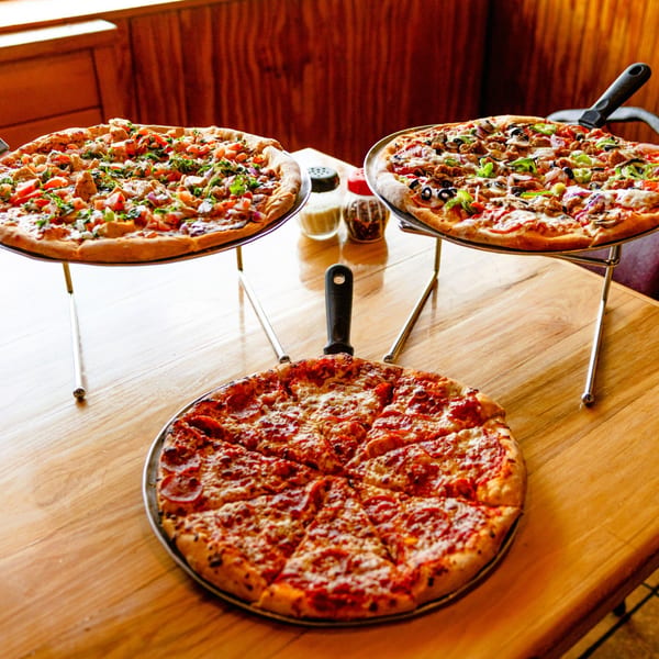 Three Halftime Pizza pizzas on stands, including pepperoni, chicken, and supreme, on a wooden table.