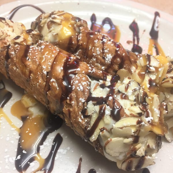 Two cannolis topped with powdered sugar, chocolate, caramel, and sliced almonds on a white plate.