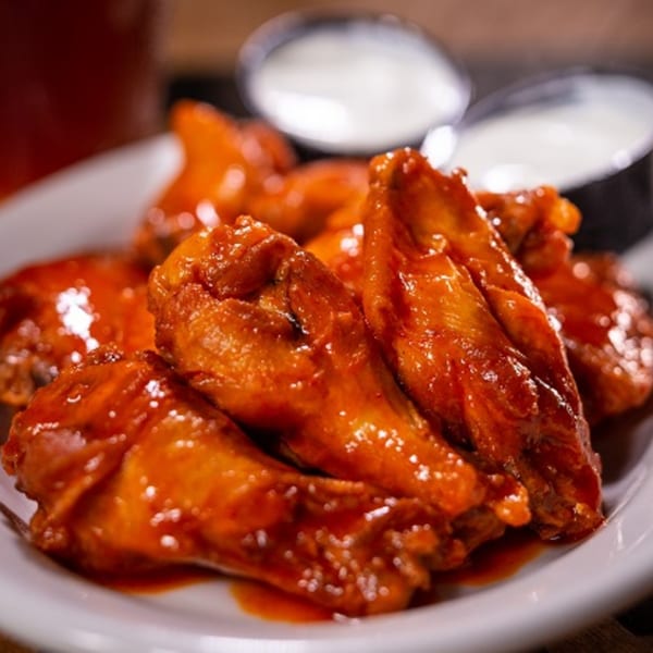 Halftime Pizza's spicy chicken wings, glistening with sauce, served on a white plate with dipping sauces.